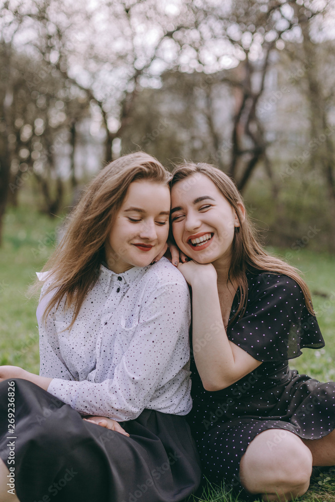 Portrait of two beautiful young sisters sitting in the green spring park, hugging in the field. Having fun together, positive emotions, bright colors. Copy space. Happy girlfriends at sunset Portrait of two beautiful young sisters sitting in the green spring park, hugging in the field. Having fun together, positive emotions, bright colors. Copy space. Happy girlfriends at sunset