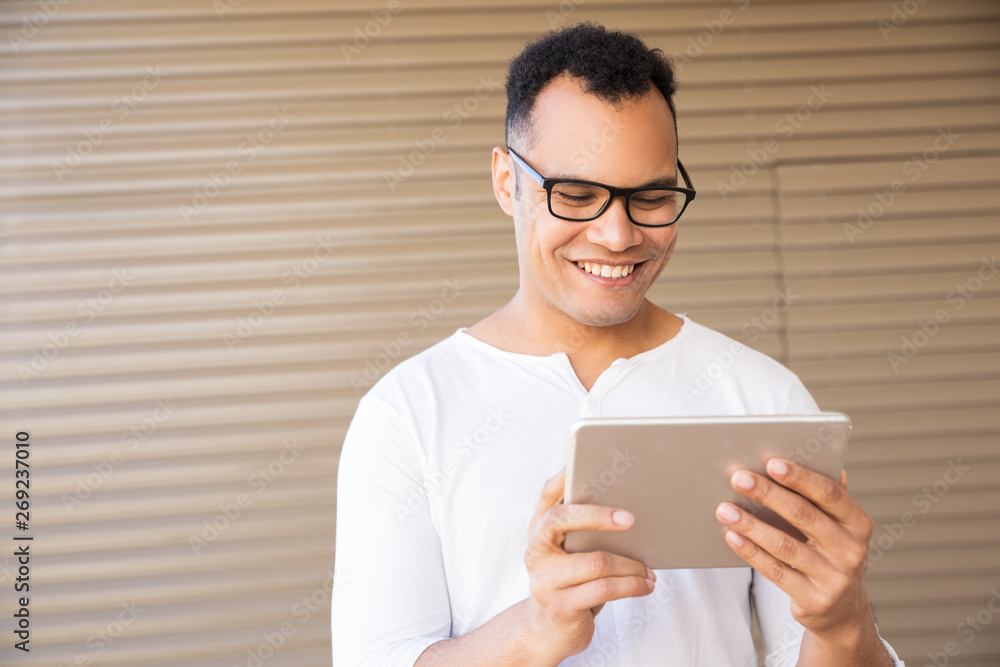 Medium shot of smiling young mixed-race man in spectacles and white T-shirt standing at office building, working on tablet. Front view. Lifestyle concept Medium shot of smiling young mixed-race man in spectacles and white T-shirt standing at office building, working on tablet. Front view. Lifestyle concept
