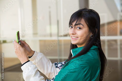 Side view of pretty young mixed-race woman in green jacket making photo on phone, turning head to camera, smiling. Lifestyle concept