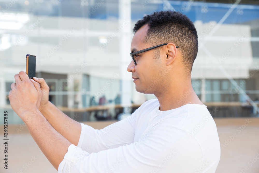 Serious young guy taking selfie photo on smartphone outdoors. Handsome man standing with building glass wall in background. Selfie photo concept. Side view. Serious young guy taking selfie photo on smartphone outdoors. Handsome man standing with building glass wall in background. Selfie photo concept. Side view.