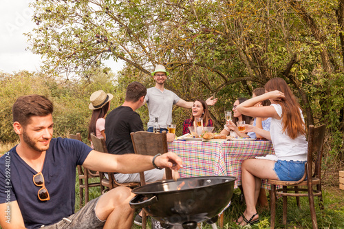 cheerful friends having fun while barbeque picnic party in the garden. guy prepares the grill others laugh and enjoy the celebration. photo