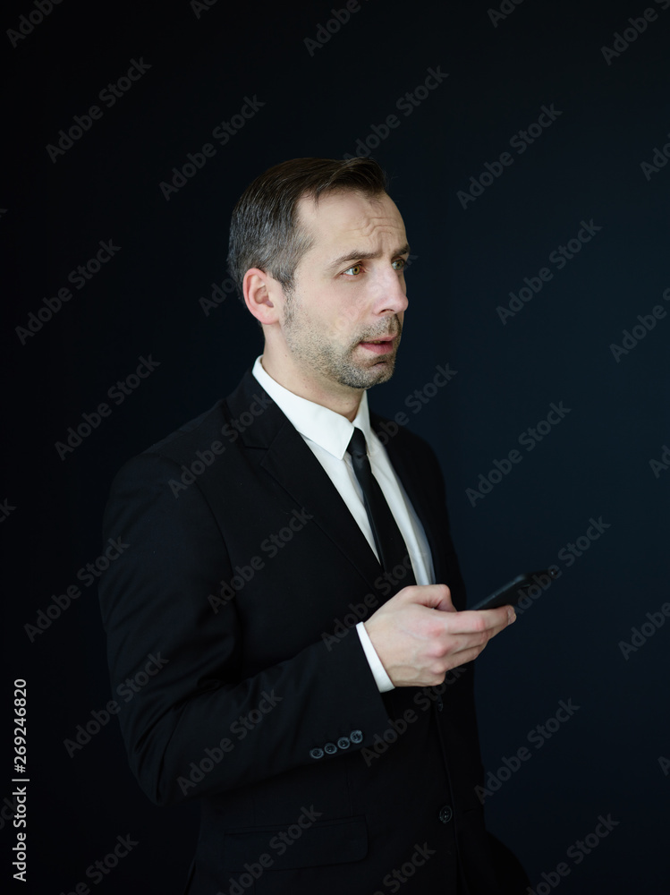 Portrait of serious middle aged businessman in formal black suit, white shirt and tie holding cell phone in his hand and looking away thoughtfully on dark background Portrait of serious middle aged businessman in formal black suit, white shirt and tie holding cell phone in his hand and looking away thoughtfully on dark background