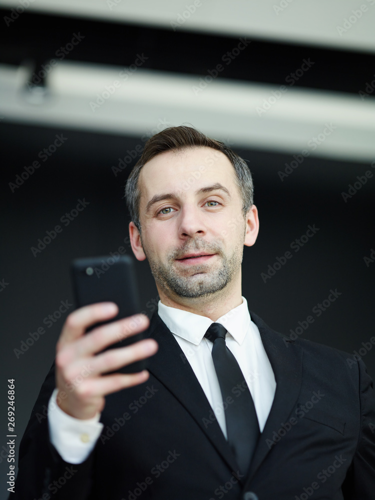 Low angle view of middle aged businessman with stubble holding smartphone and looking at camera Low angle view of middle aged businessman with stubble holding smartphone and looking at camera