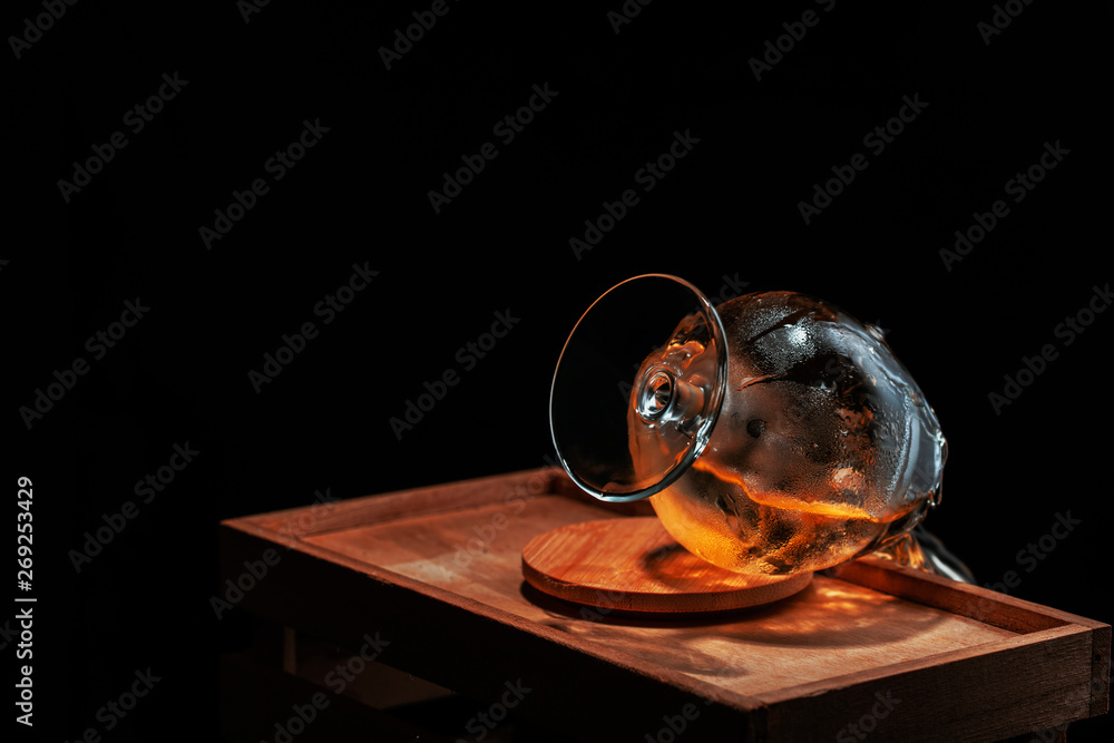  whiskey glass overturned and soaked up from cold ice from which follows drink, on a wooden support on a black background.