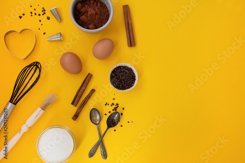 Ingredients for baking with baking utensile on yellow background - flour, wooden spoon, rolling pin, eggs, coccoa, sprinkles, banana. Top view, copy space.