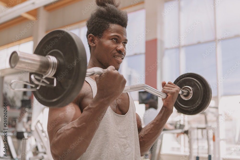 Selective focus on barbell in hands of strong African male athlete, copy space. Young muscular bodybuilder exercising at the gym, doing biceps workout Selective focus on barbell in hands of strong African male athlete, copy space. Young muscular bodybuilder exercising at the gym, doing biceps workout