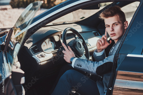 Young attractive businessman is sitting in his car at the parking while talking by mobile phone. © Fxquadro