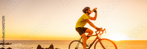 Valokuva Triahtlon athlete man drinking water bottle on road racing bike ride outdoors at sunset banner panorama landscape