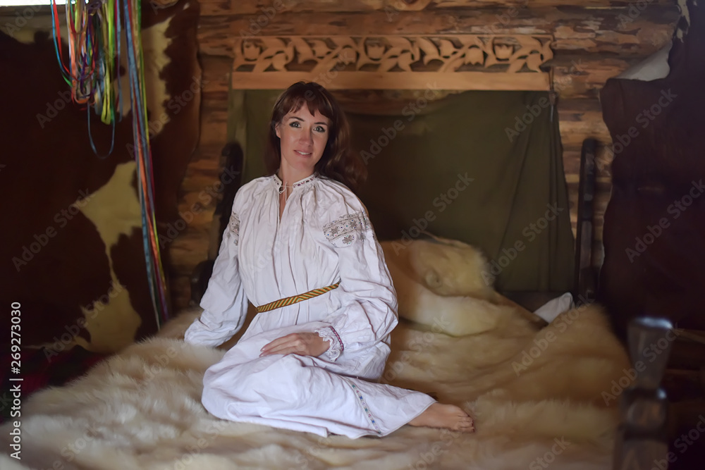 brunette in white linen old-fashioned shirt with embroidery sits on a medieval bed with a fur skin