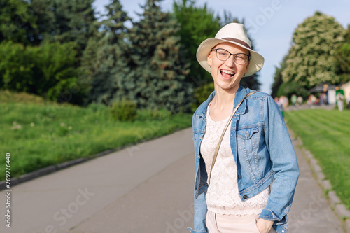 Happy young caucasian bald woman in hat and casual clothes enjoying life after surviving breast cancer. Portrait of beautiful hairless girl smiling during walk at city park after curing disease