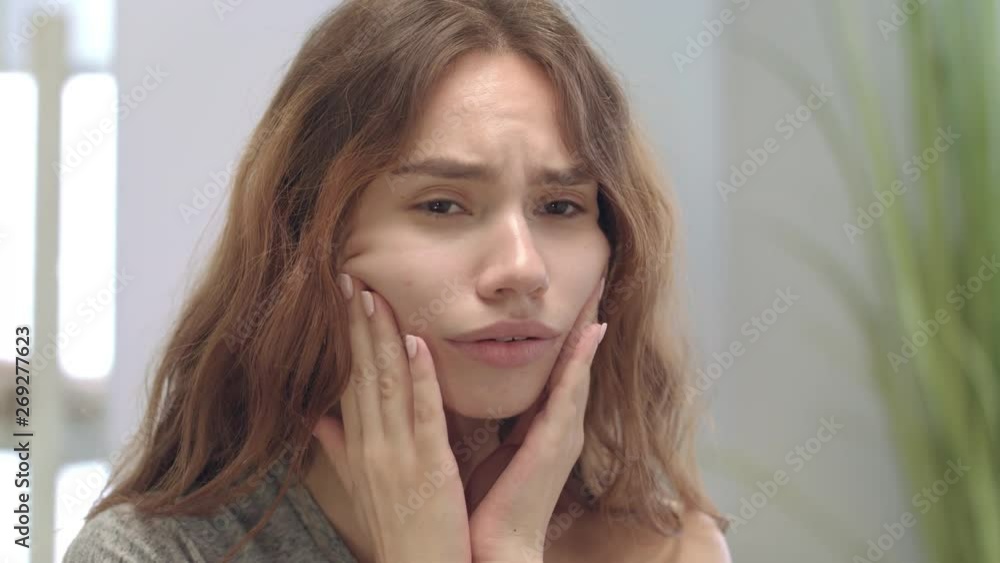 Unhappy young woman looking at face and touching skin front bathroom mirror Unhappy young woman looking at face and touching skin front bathroom mirror