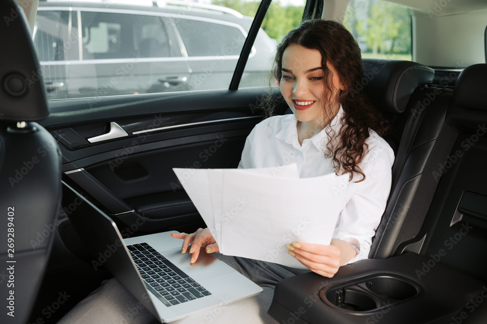 Beautiful young business woman using laptop and working with documents in the car.