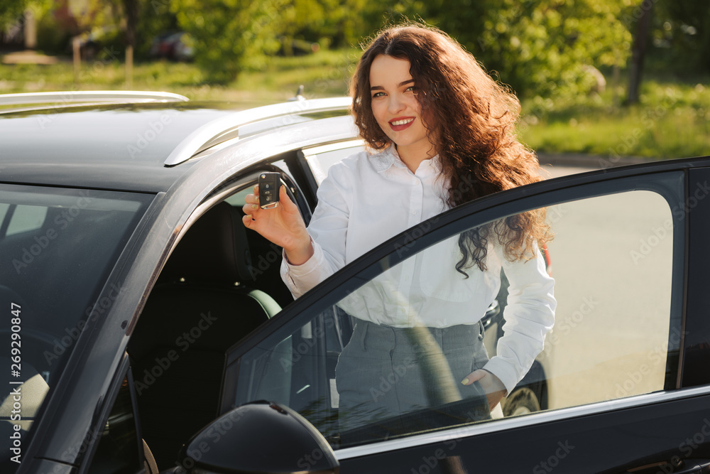 Outdoor shot of satisfied beautiful female owner of car, likes and rejoices her purchase, holds keys , has happy expression, stands near new automobile, going to drive