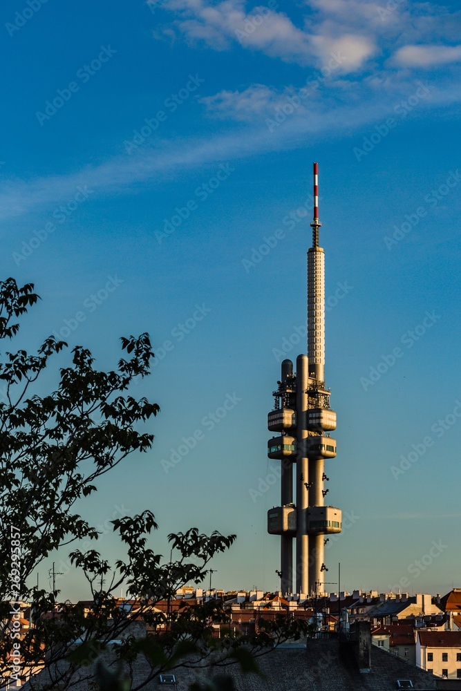 Prague, Czech Republic - May 21 2019: Aerial view of Zizkov television tower from Vitkov hill on a sunny spring evening. Blue sky with clouds. Green tree in foreground. Vertical image.