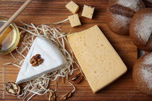 Top view of hard and brie cheeses of in assortment with nuts, honey and buns on a wooden board and light background. The concept of still life. Space for text