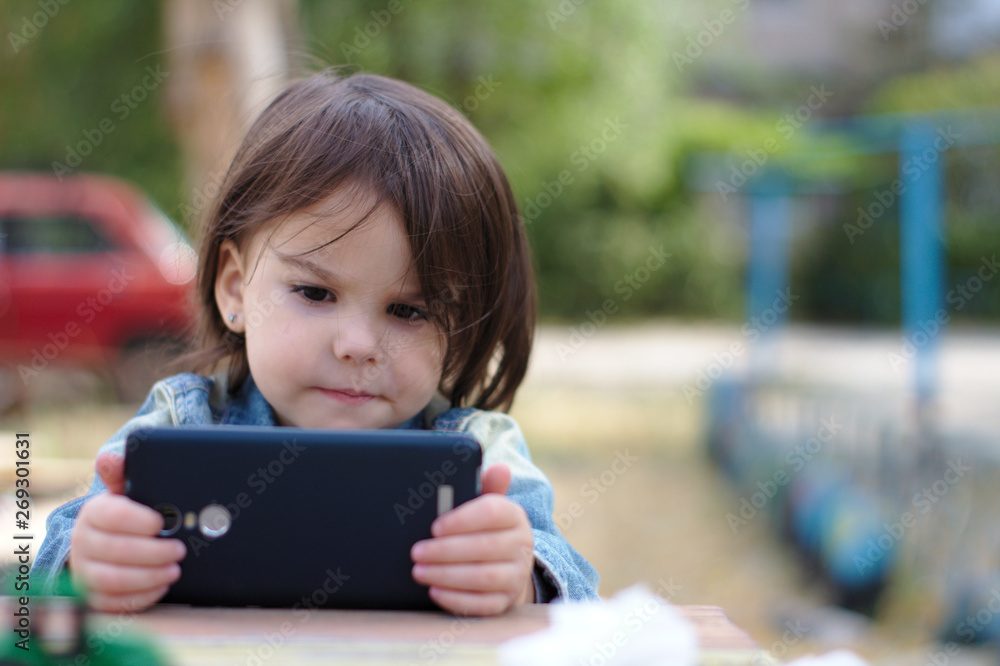 girl in a denim jacket holds in her hands and looks into the smartphone while sitting at a table on the playground girl in a denim jacket holds in her hands and looks into the smartphone while sitting at a table on the playground