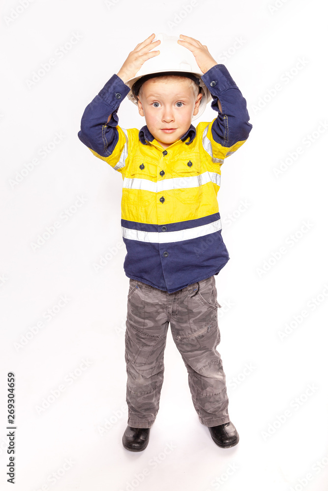 Young blond caucasian boy hands on hard hat with a shocked look role playing as a construction worker supervisor in a yellow and blue hi-viz shirt, boots. Young blond caucasian boy hands on hard hat with a shocked look role playing as a construction worker supervisor in a yellow and blue hi-viz shirt, boots.
