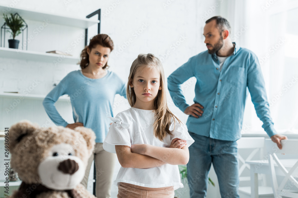 selective focus of cute kid standing with crossed arms near teddy bear and parents at home