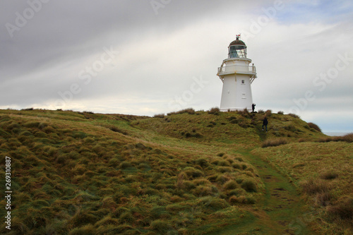 Billede på lærred White beautiful lighthouse on a green grassy hill in the cloudy day