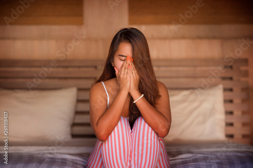 dramatic lifestyle portrait of young sad and depressed Asian Indonesian woman in pajamas sitting desperate on bed suffering pain and depression feeling lonely