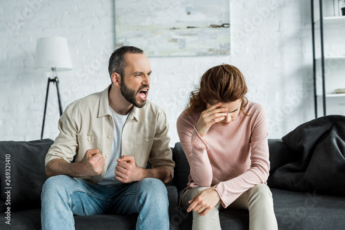 angry man sitting on sofa and screaming at upset woman