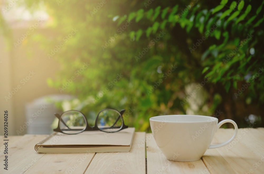 Cup of hot coffee, reading glasses and a book on wooden table in the green garden this morning. 
