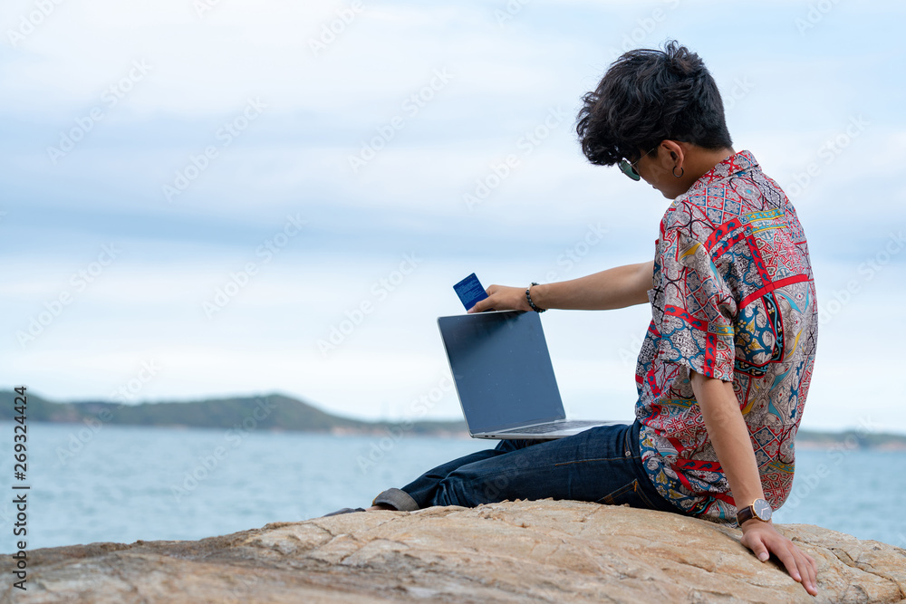 Selective focus young adult man traveler sitting on ocean cliff on tropical island in summer day vacation holding or using laptop and credit card for online shopping or working with ocean backgrounds