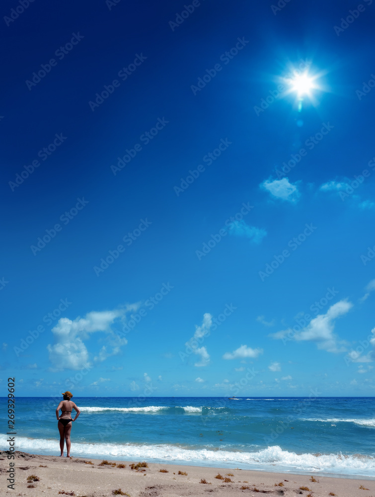 Young adult woman in bikini relaxing on sandy beach over clear blue sky with shining sun
