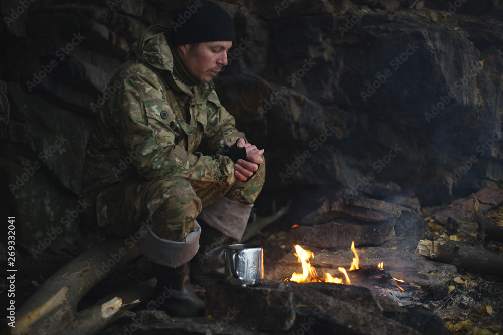 Man in camouflage drinking tea and warms himself by the fire in bad weather. Extreme travel. Survival in the wild. Man in camouflage drinking tea and warms himself by the fire in bad weather. Extreme travel. Survival in the wild.