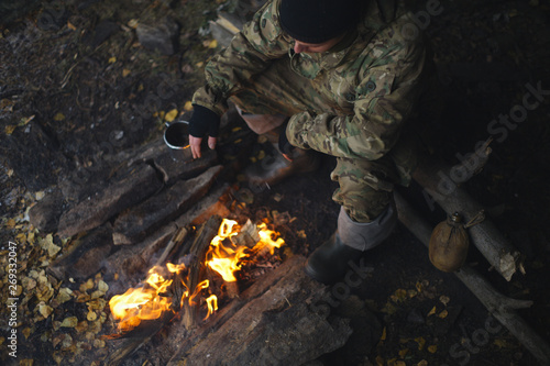 Man in camouflage drinking tea and warms himself by the fire in bad weather. Extreme travel. Survival in the wild.