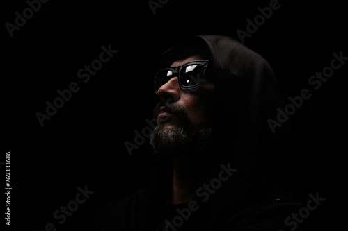 Brutal man with grey beard in sunglass with hood on dark background.