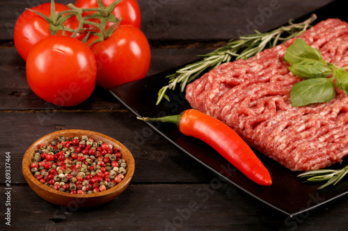 raw minced meat on paper, ingredients for burger with tomato, pepper, onion, spices and seasonings on black background