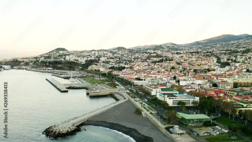 An aerial cityscape view of Funchal, Madeira, showing traffic, Harbour & buildings along coastal city with the mountain range in the distance. An aerial cityscape view of Funchal, Madeira, showing traffic, Harbour & buildings along coastal city with the mountain range in the distance.