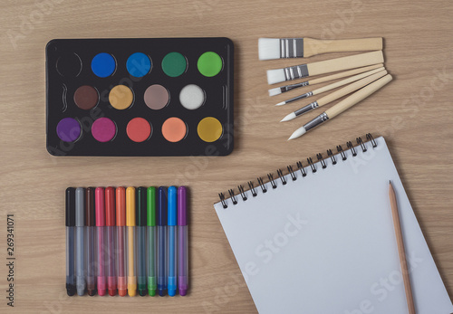 Notepad or notebook with Many colorful pens, paintbrush and Watercolor palette on brown wood table.using for Arts and Education background