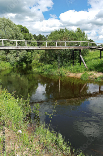 Old wooden bridge over a small river. Reflection of blue sky and white clouds in the water of a forest river. On the banks of the river grow large green trees. Summer day.