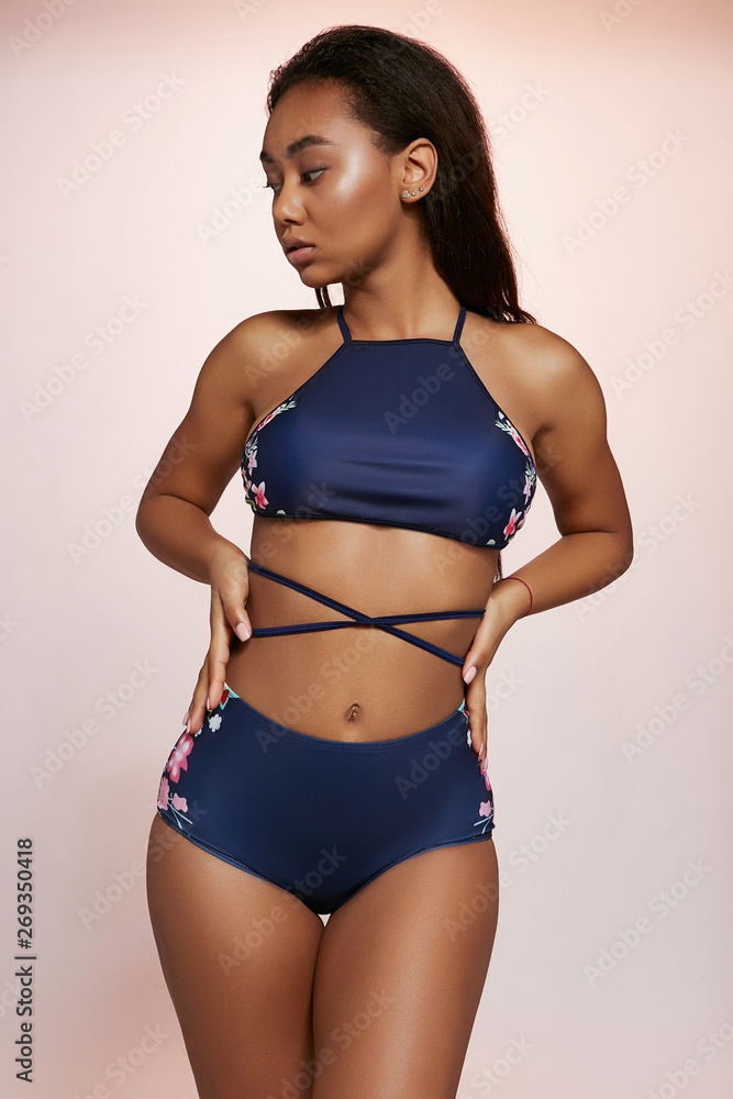 Fototapeta premium Three quarter shot of African lady, wearing navy blue two-piece swimsuit with criss-crossed strings and floral pattern. The girl is turning head to side, with hands on waist on the creamy background.