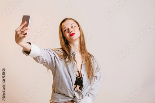 Happy cute girl in a light casual clothing makes selfie, looks at the smartphone and smiles. Cute blonde hair and red lips woman makes a photo on the phone.