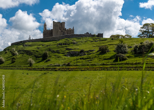 A view across green fields to the magnificent Rock of Cashel and the stone fortress and abbey, against a bright blue sky and fluffy white clouds, nobody in the image