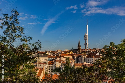Prague, Czech Republic - May 21 2019: Aerial view of Zizkov television tower and gothic church of St. Prokop from Vitkov hill on a sunny spring evening with blue sky. Green tree in foreground.