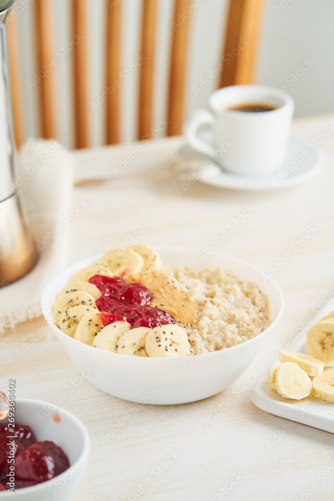 Oatmeal porridge with strawberry jam, peanut butter, banana, Chia on white wooden light background. Healthy vegan diet breakfast. Vertical. Oatmeal porridge with strawberry jam, peanut butter, banana, Chia on white wooden light background. Healthy vegan diet breakfast. Vertical.