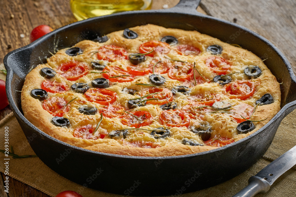 Focaccia, pizza in skillet. Close up italian flat bread with tomatoes, olives and rosemary. Side view, wooden table