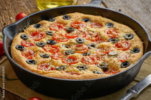 Focaccia, pizza in skillet. Close up italian flat bread with tomatoes, olives and rosemary. Side view, wooden table