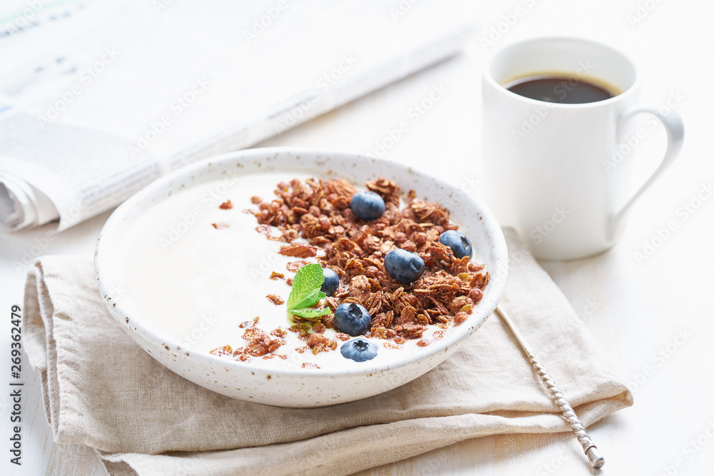 Breakfast with cup of coffee, newspaper, yogurt with chocolate granola, bilberry on a white background, side view Breakfast with cup of coffee, newspaper, yogurt with chocolate granola, bilberry on a white background, side view