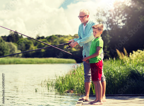family, generation, summer holidays and people concept - happy grandfather and grandson with fishing rods on river berth