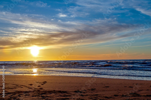 Sand beach with endless horizon and foamy waves under the bright sundown with yellow colors and clouds above the sea