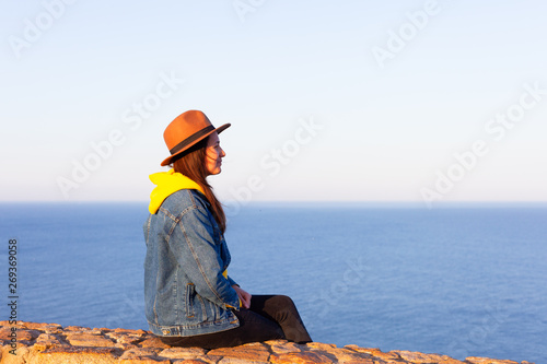 Woman in travel clothes and hat sitting and looking at blue ocean and sky.