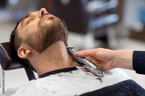 grooming and people concept - man and barber with trimmer or shaver cutting beard at barbershop