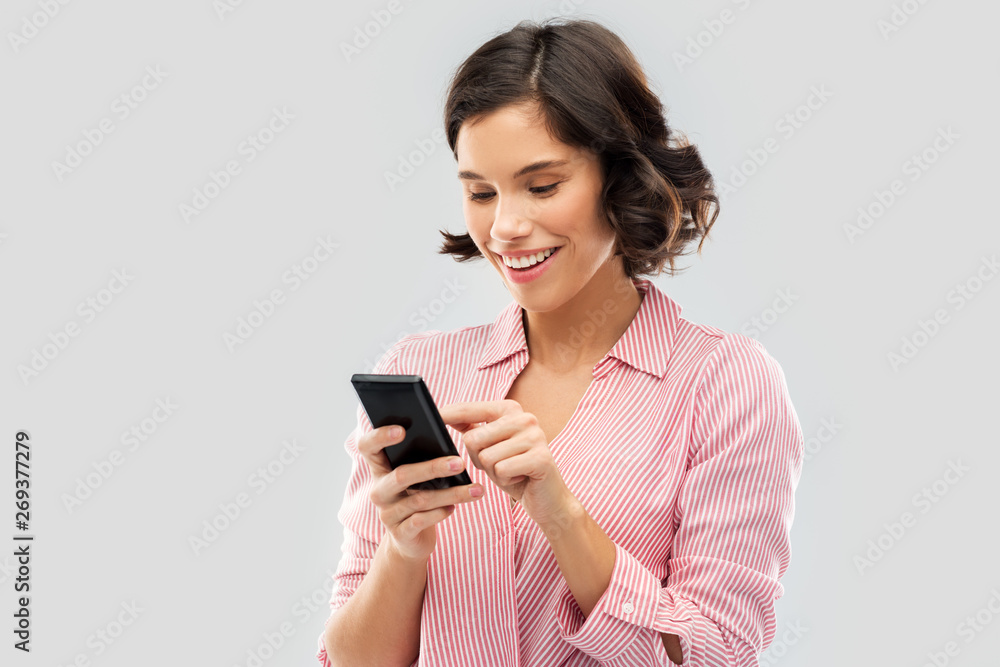technology and people concept - happy smiling young woman in striped shirt using smartphone over grey background technology and people concept - happy smiling young woman in striped shirt using smartphone over grey background