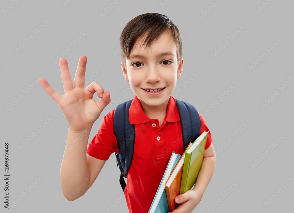 school, education and people concept - smiling little student boy in red polo t-shirt in glasses with books and bag showing ok hand sign over grey background school, education and people concept - smiling little student boy in red polo t-shirt in glasses with books and bag showing ok hand sign over grey background
