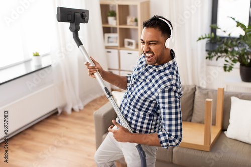 household and cleaning concept - indian man in headphones with vacuum cleaner having fun at home photo